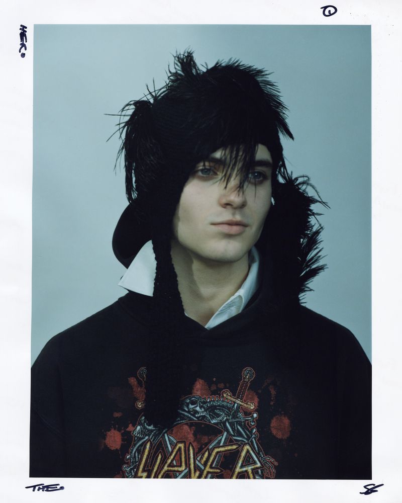Portrait of a young adult wearing a black hoodie with graphic print and a unique feathered hat against a neutral background.