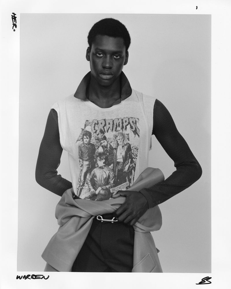 Monochrome portrait of a Black male model wearing a graphic t-shirt, dark trousers and a light jacket wrapped around the waist in studio lighting.