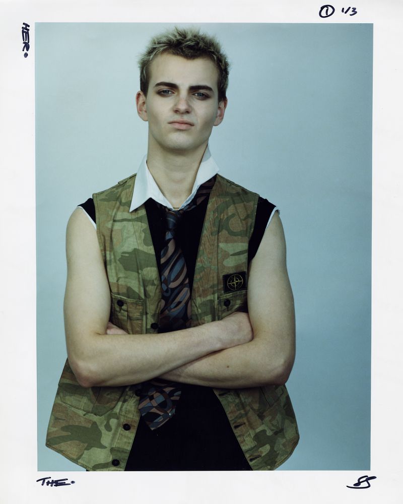 Portrait of a young adult with light hair wearing a camouflage vest, black sleeveless shirt, tie, and heavy makeup against a light grey backdrop.