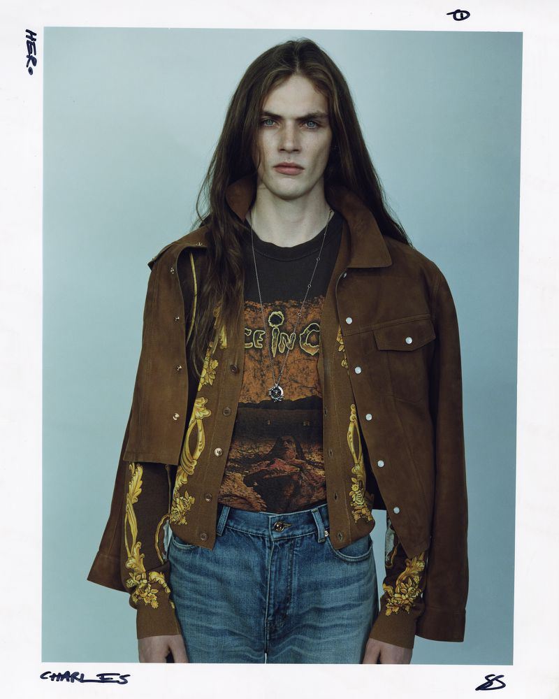 A young adult male with long brown hair, wearing a brown jacket and graphic tee, is posing indoors against a neutral light-blue wall.