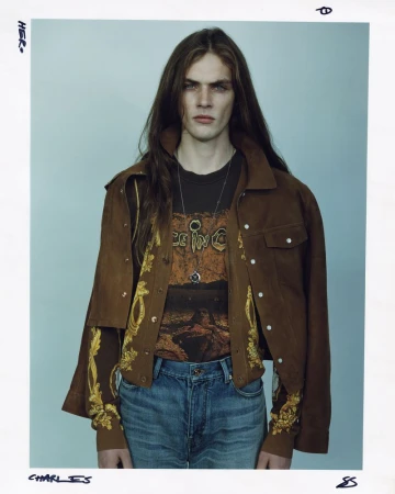 A young adult male with long brown hair, wearing a brown jacket and graphic tee, is posing indoors against a neutral light-blue wall.
