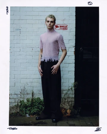 A young male model with bleached hair wearing a light pink sheer knit top and black trousers standing against a white brick wall.