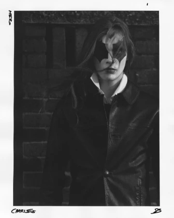Monochrome portrait of a model wearing a dark leather jacket with bat-wing eye makeup standing before a brick wall.