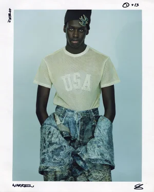 Young Black man wearing a mesh USA t-shirt, beanie, and patterned denim jeans against a light background