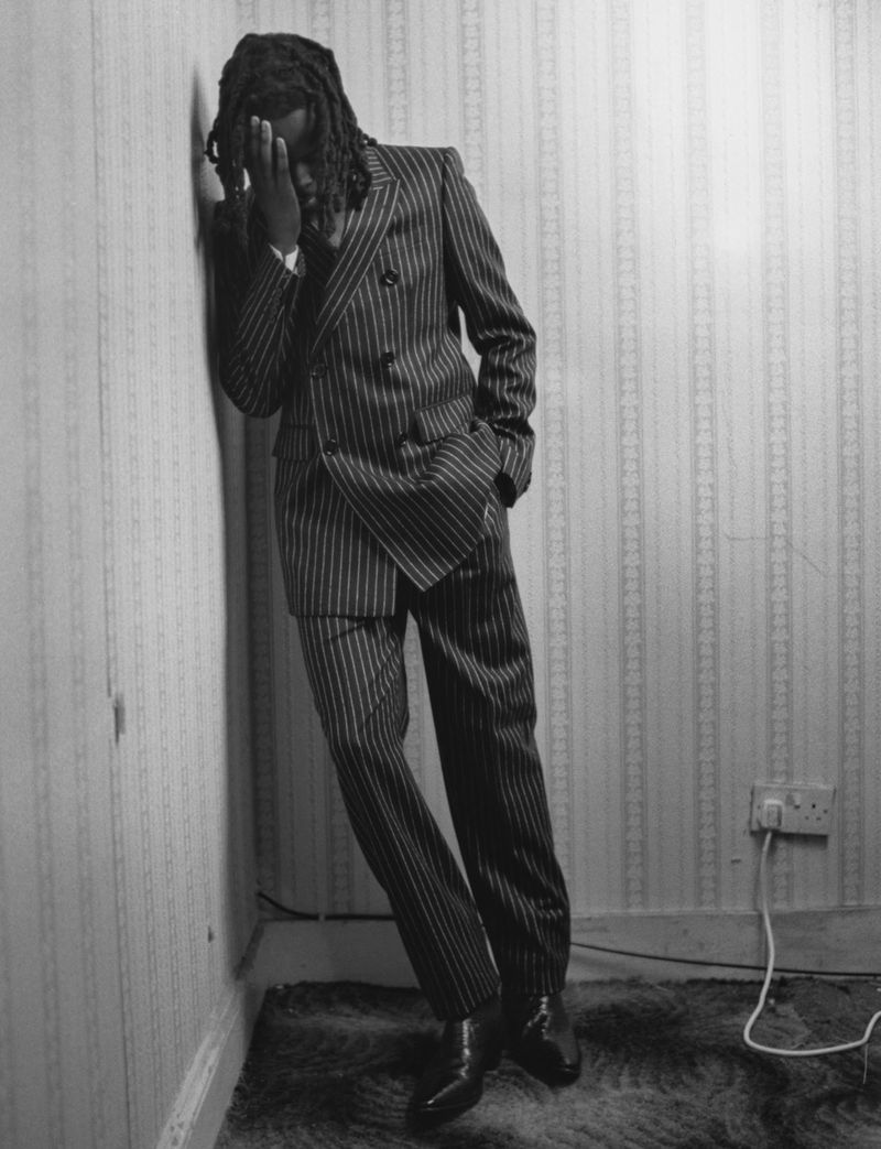 Black and white portrait of a man with dreadlocks wearing a pinstripe suit leaning against a striped wall in an indoor setting.
