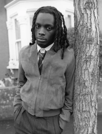 Black man with dreadlocks wearing a suede jacket and striped tie leaning against a tree trunk outdoors in a black and white portrait.