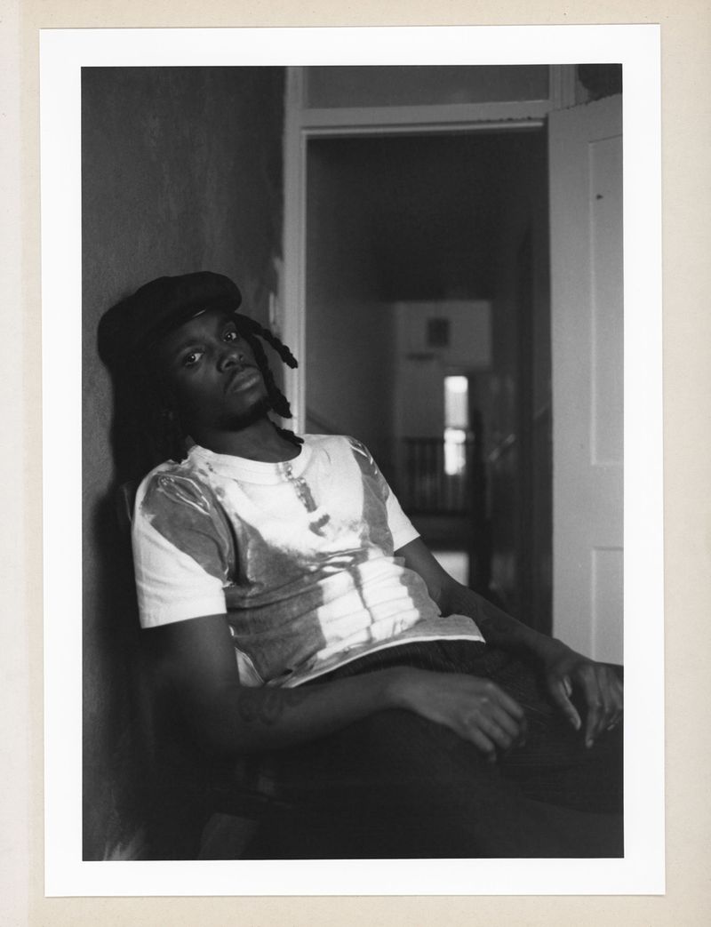Monochrome portrait of a young adult male with dreadlocks wearing a beret and a printed t-shirt leaning against a wall indoors.