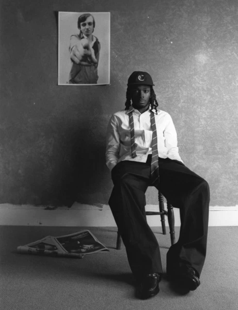 Monochromatic portrait of a young adult seated, wearing a baseball cap and tie, with a newspaper and poster in an indoor setting.