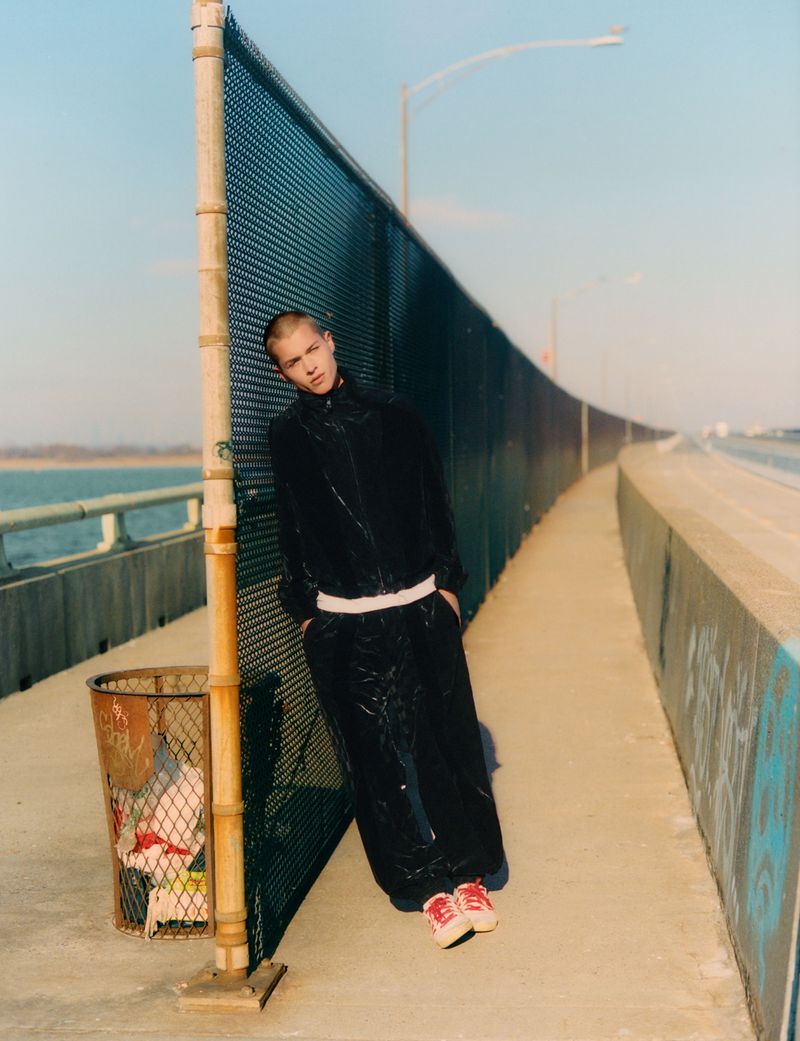 Young man in dark tracksuit leans against fence on urban bridge under bright sunlight, overlooking distant shoreline and street lights.