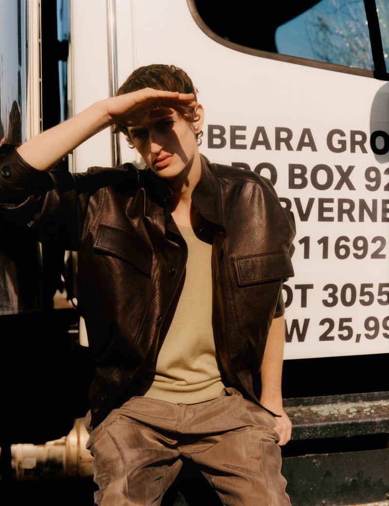 Male model in a brown leather jacket and matching pants shields his eyes by an inscription covered truck on a sunny day outdoors.