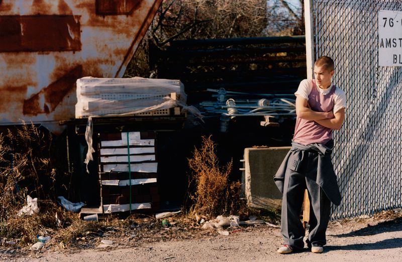 Young adult stands with arms crossed by a metal fence near an old trailer and overgrown foliage in a gritty outdoor setting.