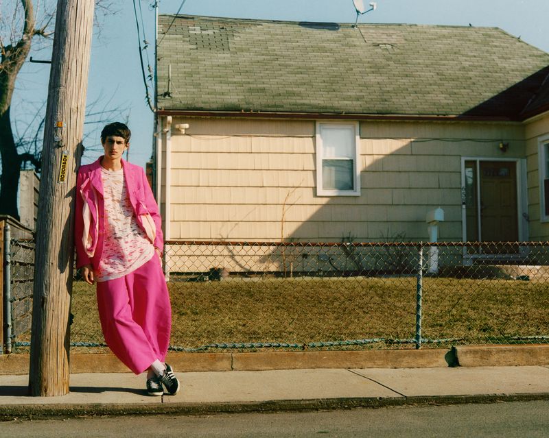 A young man poses in bright pink clothing, leaning casually against a pole outside a home on a sunny day.