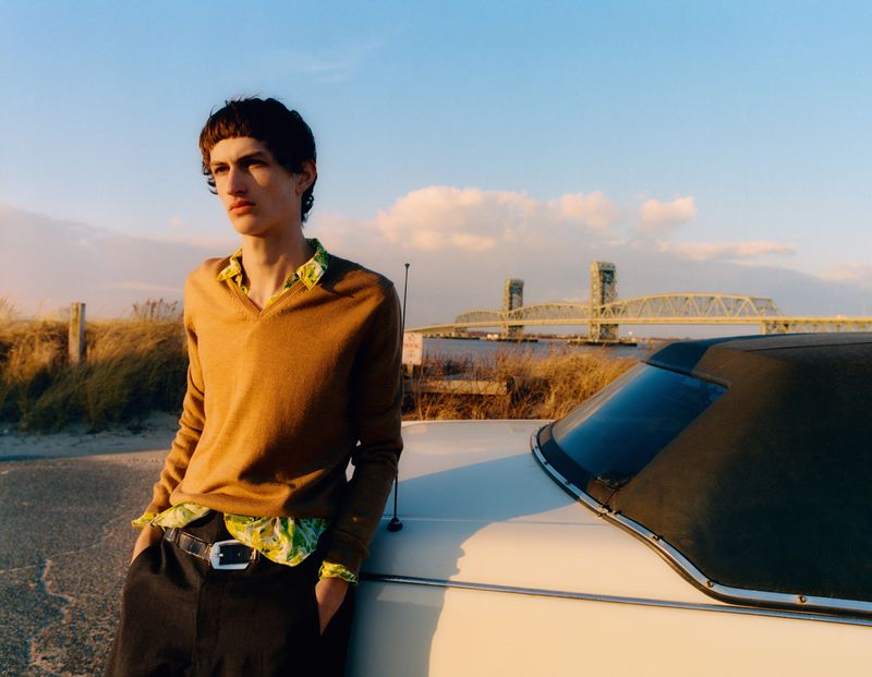 Young man in a brown sweater leans against a vintage car on a beach with a bridge in the background, shot at eye-level in the daytime.