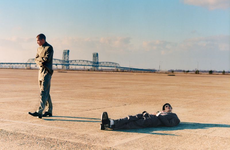 Two adult men pose outdoors on a barren landscape with a bridge in the distance on a sunny day, wearing similar colored jackets and pants.