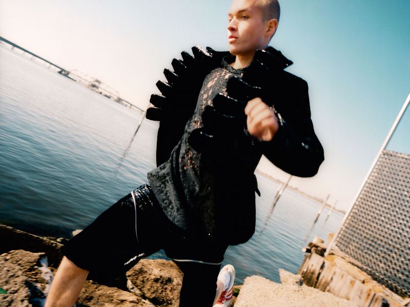 Man in a black jacket stands on rocks by the water with a bridge visible in the background on a sunny day in a fashion shot.