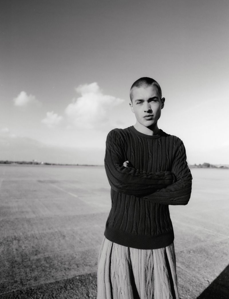 Monochrome fashion portrait of a young man with a shaved head wearing a dark textured sweater outdoors.