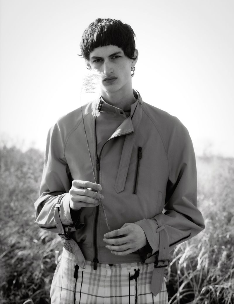 Monochrome portrait of a young adult male model in a jacket and plaid pants, standing in a field, holding a stem of wheat.