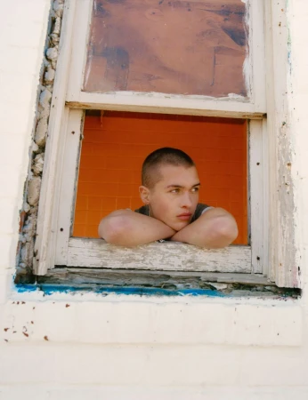 Portrait of a young adult male with short hair leaning on a weathered window frame with an orange tiled interior in the background.