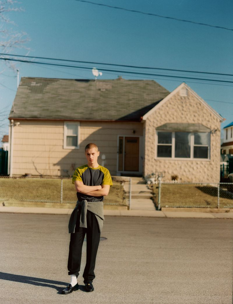 Young man poses confidently on an empty street in front of a small, quaint one-story house on a sunny day. Casual fashion style.