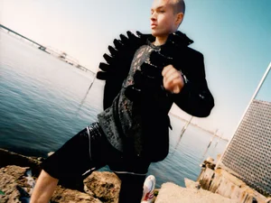 Model in black jacket with ruffled details and dark patterned shirt standing outdoors near water for HERO Magazine.