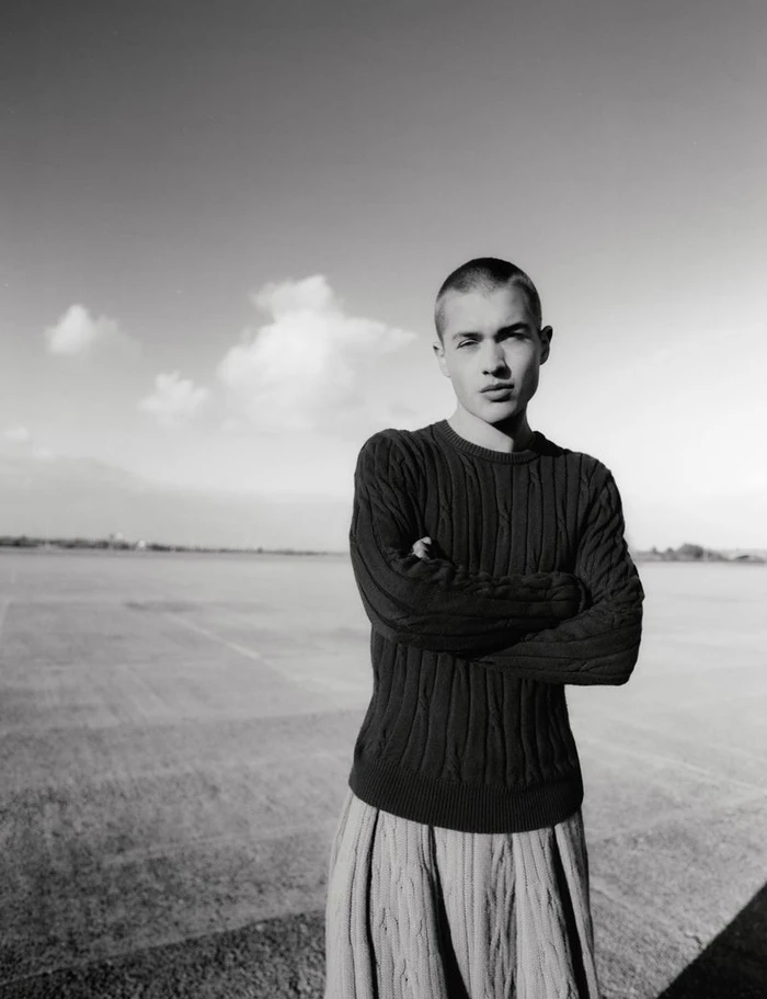 Monochrome fashion portrait of a young man with a shaved head wearing a dark textured sweater outdoors.