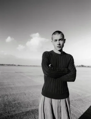 Monochrome fashion portrait of a young man with a shaved head wearing a dark textured sweater outdoors.