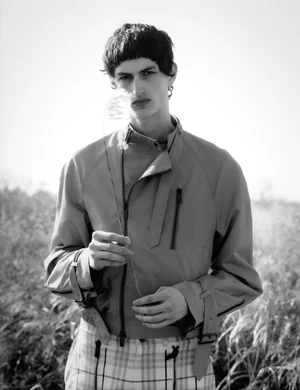 Monochrome portrait of a male model holding a thin stalk of grass outdoors in a grassy field wearing a jacket.