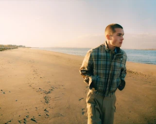 Young male model walking along a sandy beach wearing a plaid jacket and trousers during golden hour