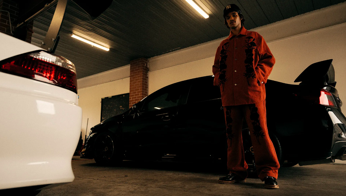 An adult male poses near two modified tuner cars in a garage setting, wearing a patterned rust-colored suit and a baseball cap.