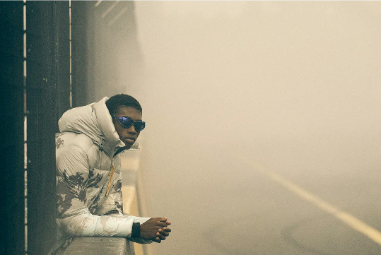 An adult male leans on a concrete barrier. He is wearing sunglasses and a hooded puffer jacket with a misty background. 