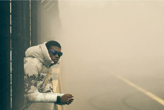 An adult male leans on a concrete barrier. He is wearing sunglasses and a hooded puffer jacket with a misty background. 