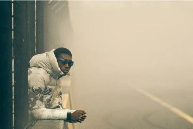 Black man wearing sunglasses and a patterned puffer jacket leaning on a bridge rail in heavy fog