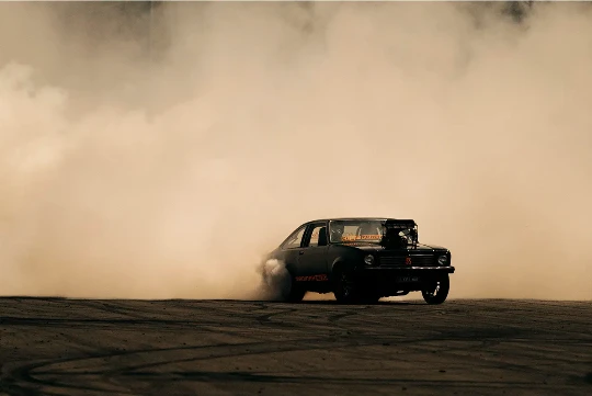 Vintage black car drifting and leaving tire tracks on the ground, surrounded by a cloud of smoke in an outdoor environment.
