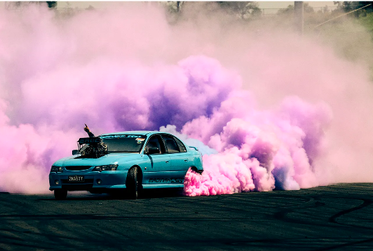 Light blue car performs a burnout, creating a cloud of pink and purple smoke in a dynamic outdoor automotive shot.