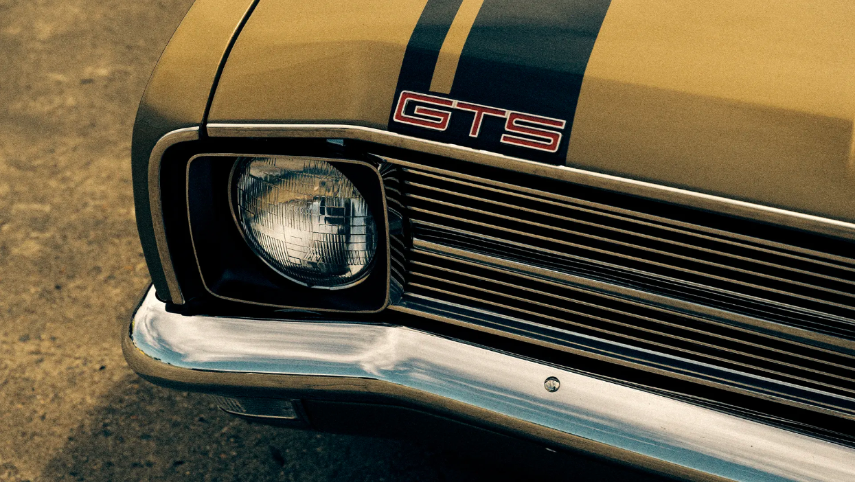 Close-up of a classic gold car with black racing stripes, showing the headlight, grill, chrome bumper, and 'GTS' emblem.