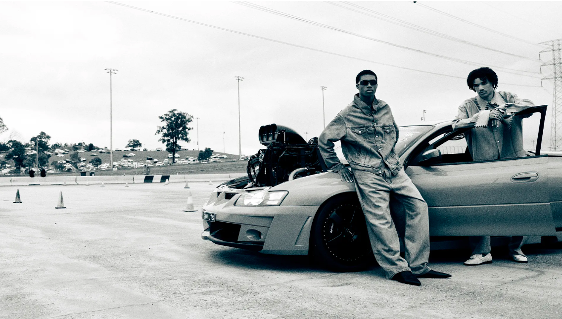 Two men in denim pose with a customized car with its hood open on an outdoor racetrack, featuring a visible engine.