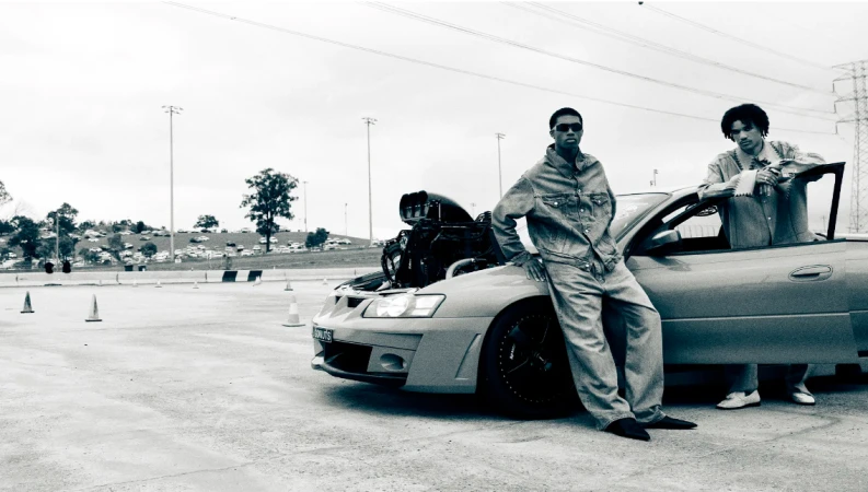 Two men in denim pose with a customized car with its hood open on an outdoor racetrack, featuring a visible engine.