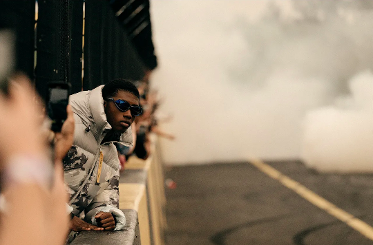 A young Black man wearing sunglasses and a patterned puffer jacket leaning over a barrier amid smoke.