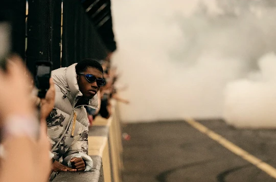 A young Black man wearing sunglasses and a patterned puffer jacket leaning over a barrier amid smoke.