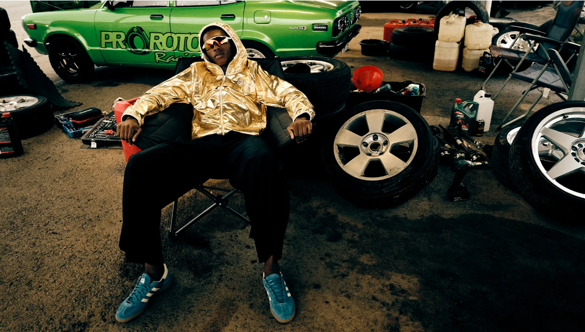 Adult male in metallic jacket and sunglasses sits outdoors surrounded by car tires and tools with a green race car in the background.