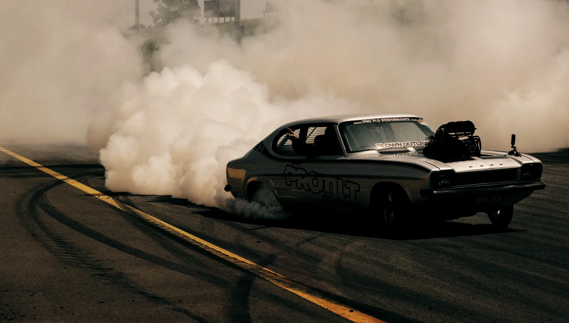 A vintage silver race car performing a burnout on an asphalt track surrounded by a cloud of white smoke with yellow lane markings.
