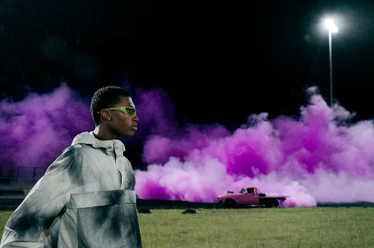 A young black man stands next to a pink truck surrounded by violet smoke in a nighttime outdoor setting with a stadium light.