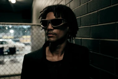 Man wearing dark sunglasses and a black jacket posing against a dark brick wall with a chain-link fence in the background.