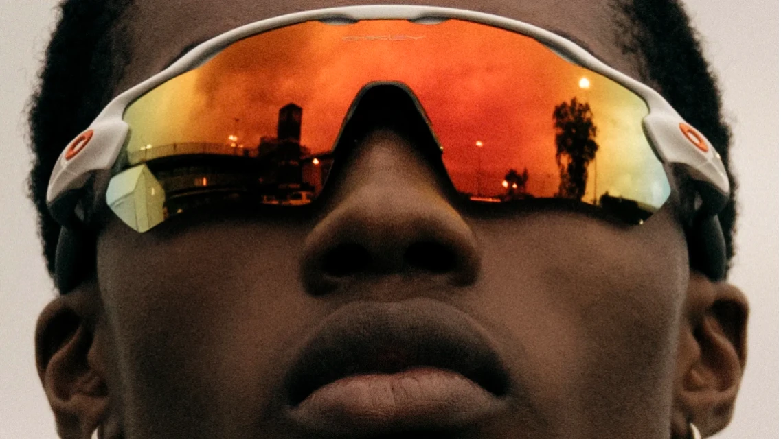 Close-up of Black adult wearing mirrored sunglasses reflecting a cityscape with buildings and streetlights against an orange sky.