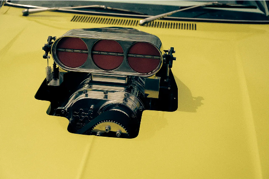 Close-up of a yellow vintage car hood featuring a prominent chrome-plated engine supercharger with three red-colored air intakes.