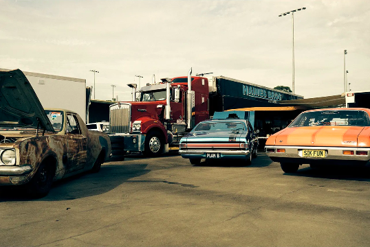 Classic cars and a semi-truck are parked in an outdoor lot on a sunny day, featuring vintage vehicles and commercial transport.