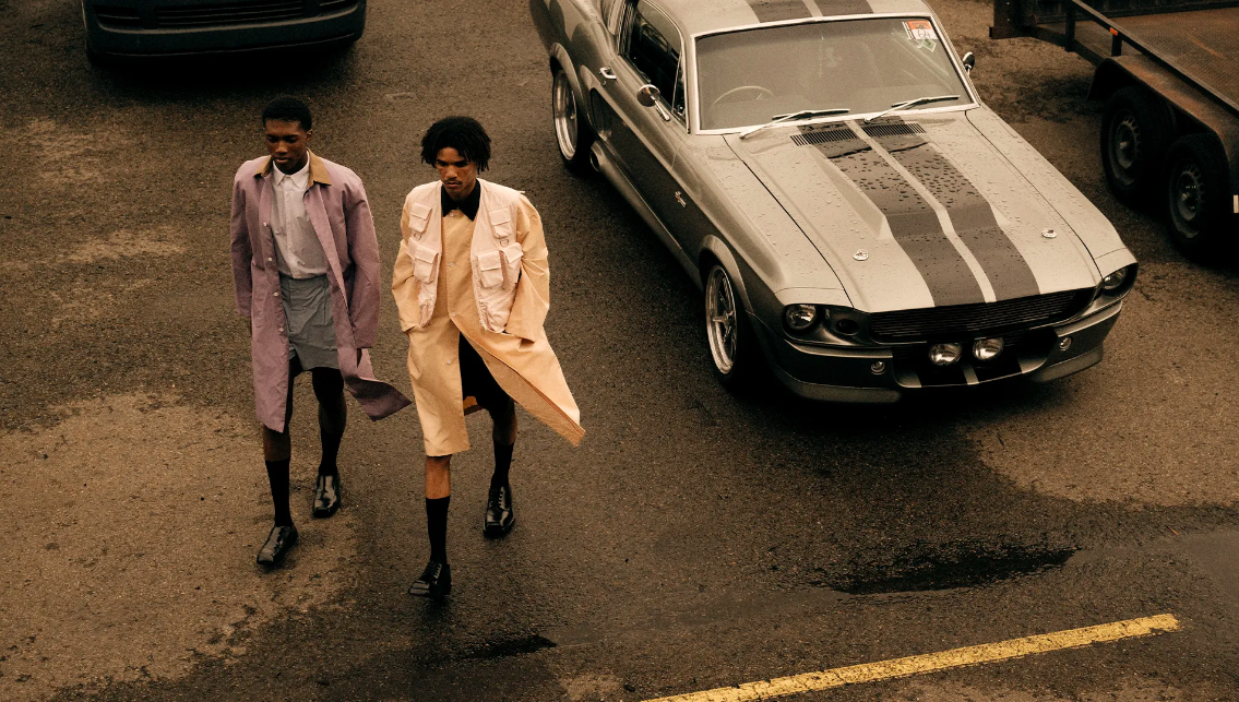 Two African American men in long coats walk next to a vintage car in an outdoor scene with asphalt ground and overcast lighting.