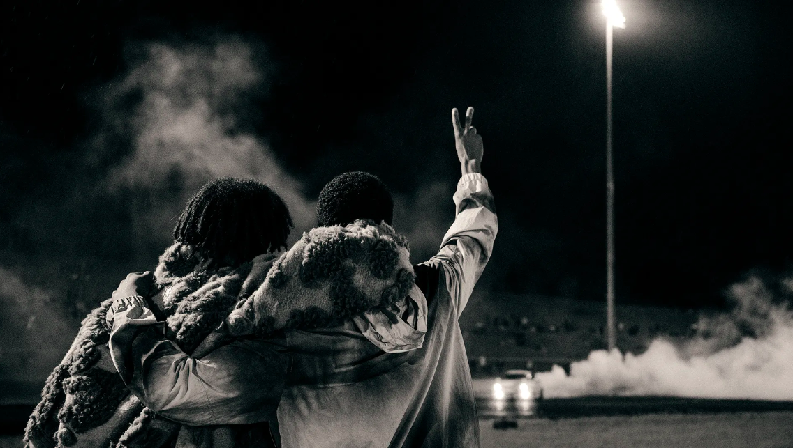 Two people embracing outdoors at night with a street light and car headlights visible in the background, captured in black and white.