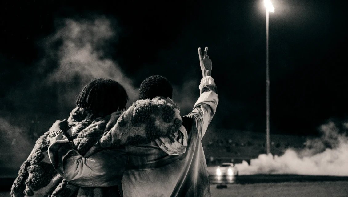 Two people embracing outdoors at night with a street light and car headlights visible in the background, captured in black and white.