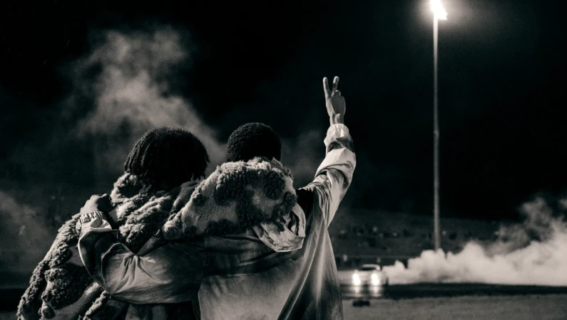 Two people embracing outdoors at night with a street light and car headlights visible in the background, captured in black and white.